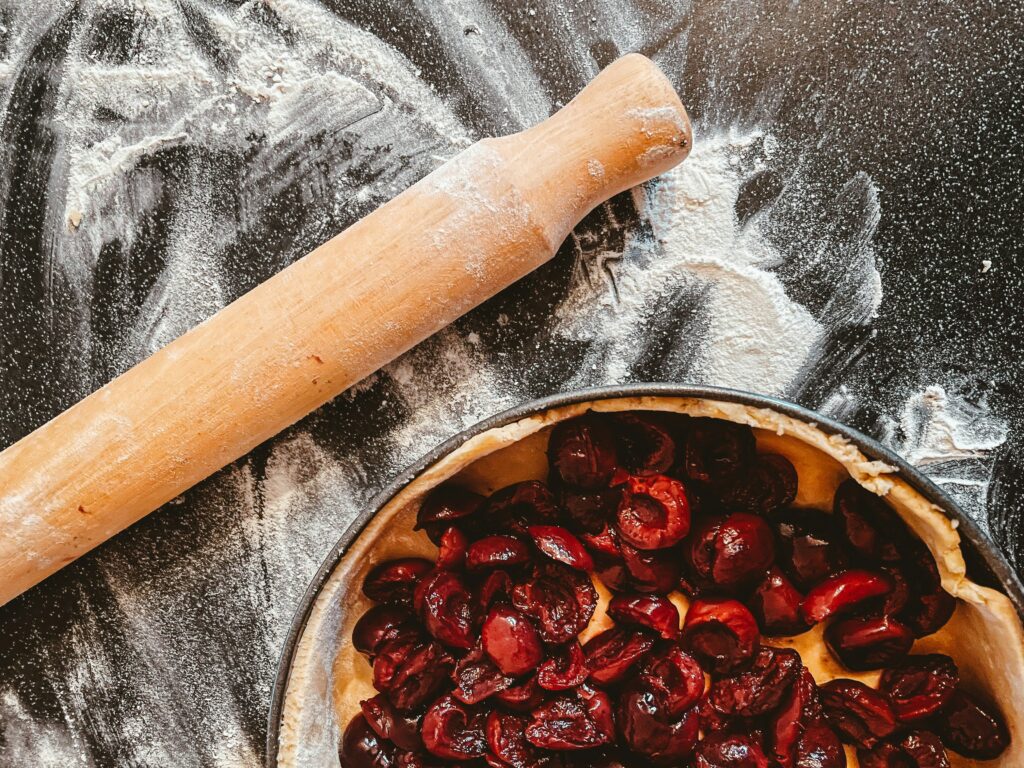 A rolling pin on a floured surface, next to a pie tin containing crust and pitted halved cherries. Photo by Nik. https://unsplash.com/@helloimnik